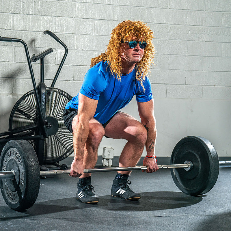 Een man met een grote bruine krulpruik en een zwarte zonnebril met blauwe reflecterende glazen tilt een zware halter op in een sportschool.