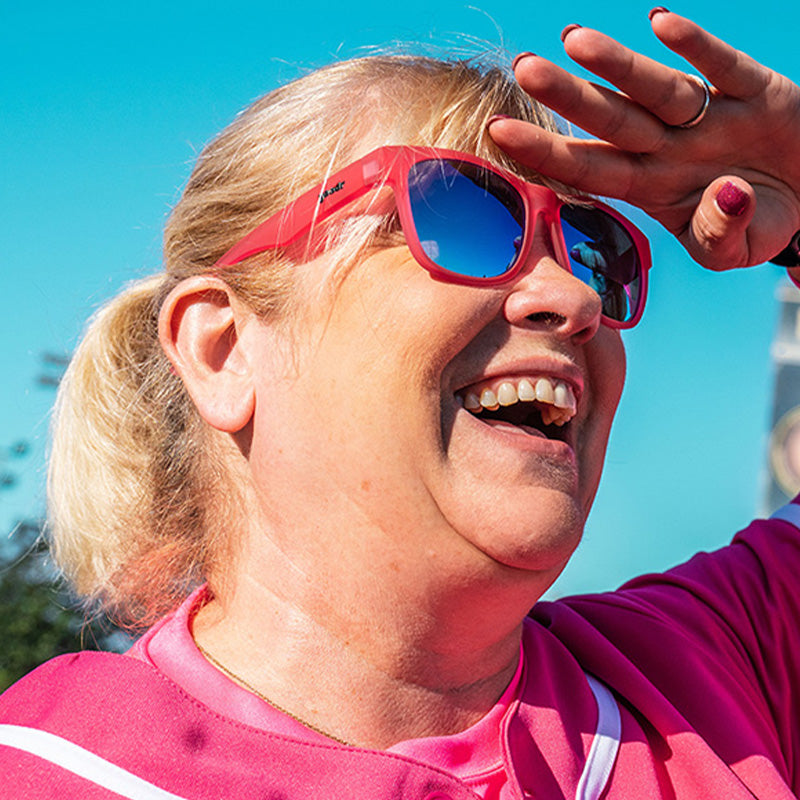 Driekwart bovenaanzicht van een dame in een roze honkbaluniform en een roze zonnebril met blauwe glazen die glimlacht op een honkbalveld.