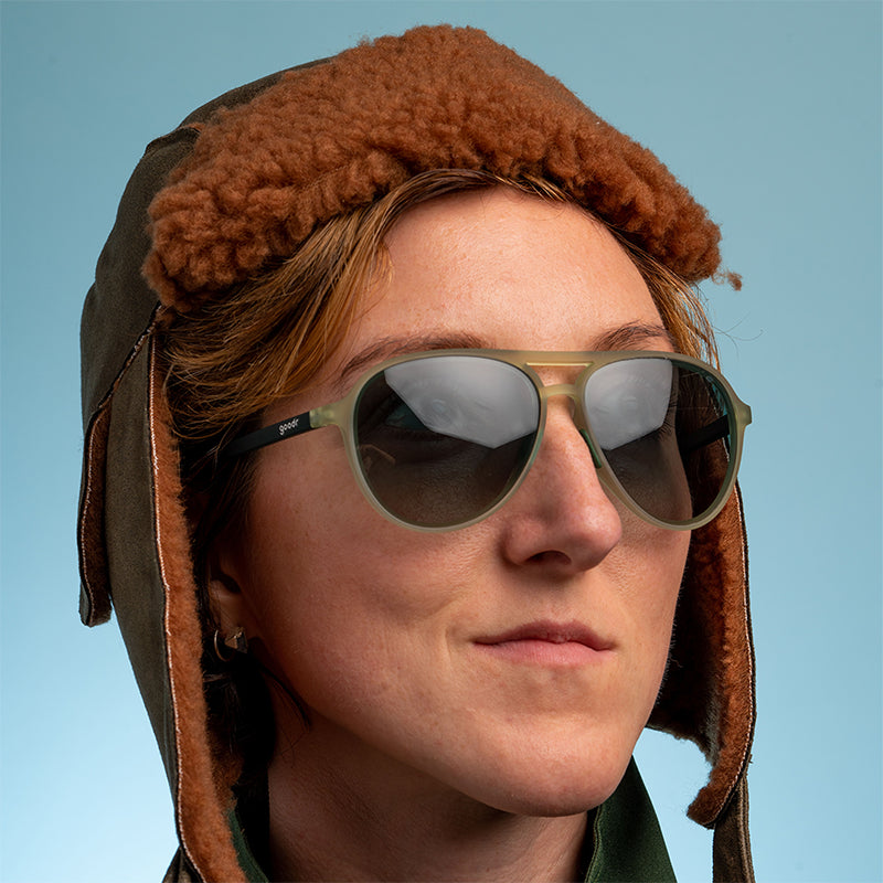 Een vrouw met een bruine, pluizige vliegenierspet kijkt zelfverzekerd naar buiten en draagt cadetgroene vliegeniers met glazen met een groen kleurverloop.