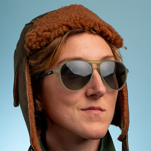Een vrouw met een bruine, pluizige vliegenierspet kijkt zelfverzekerd naar buiten en draagt cadetgroene vliegeniers met glazen met een groen kleurverloop.