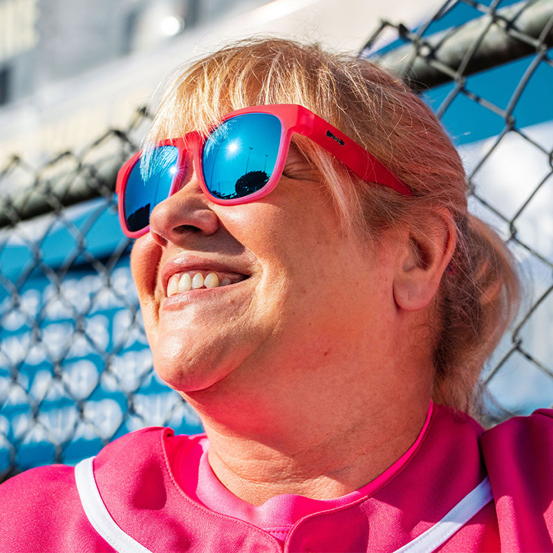 Een vrouw in een roze honkbaluniform en een roze zonnebril met blauwe reflecterende glazen glimlacht, leunend tegen een hek.
