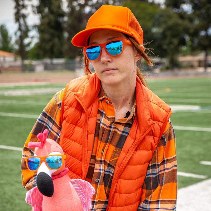 Een vrouw in een fel oranje outfit houdt een opgezette roze flamingo vast. Beiden dragen een oranje zonnebril met blauwe glazen.