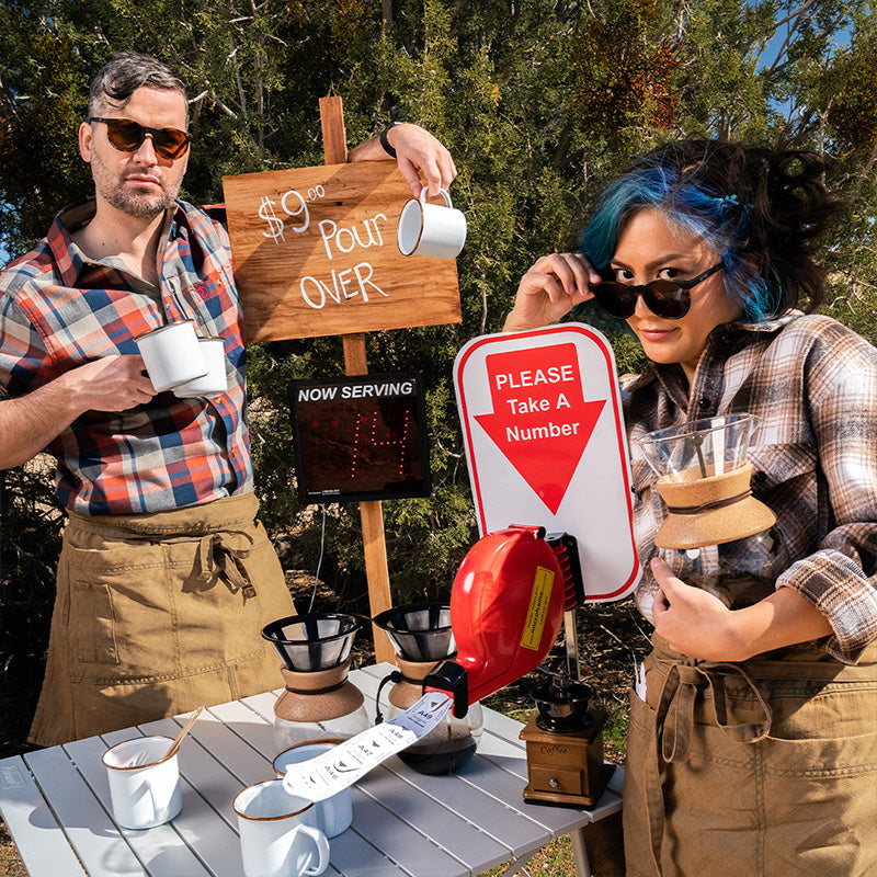 Twee hipster barista's met ronde bruine zonnebrillen met bruine glazen poseren bij een stand met ambachtelijk geschonken koffie.