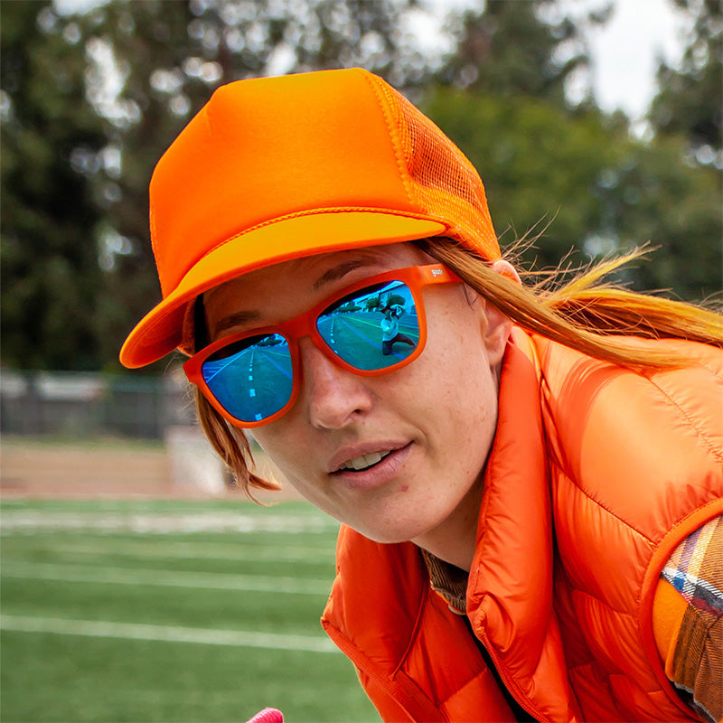 Een vrouw in een feloranje outfit staat op een voetbalveld en draagt een feloranje zonnebril met blauwe spiegelende glazen.