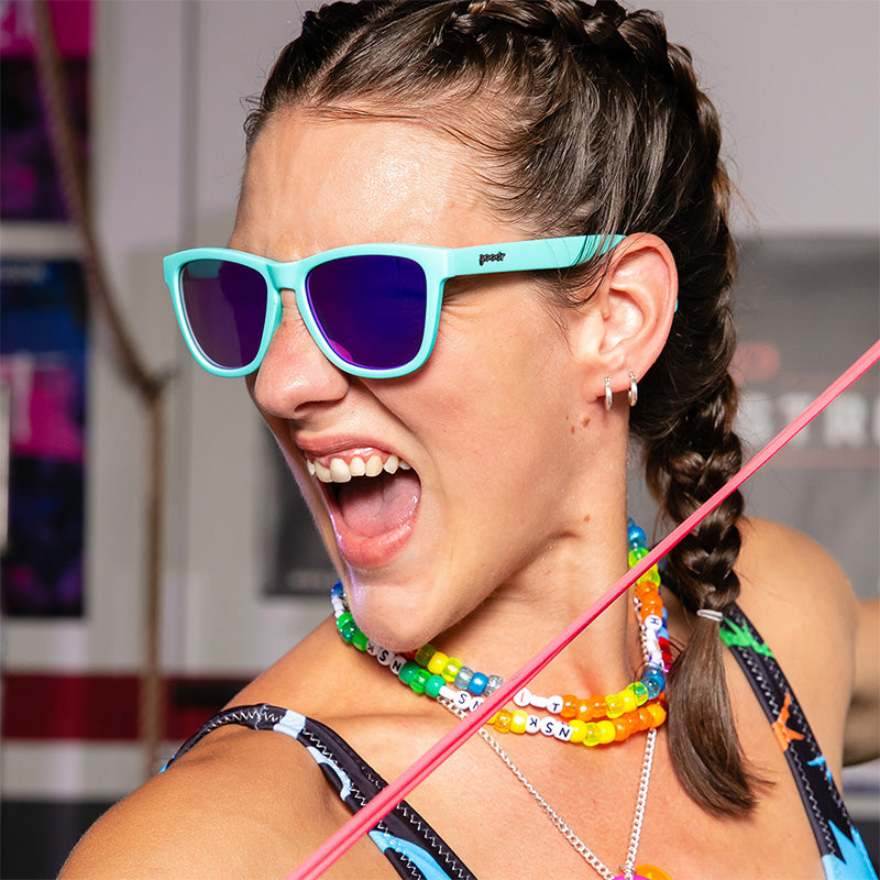 Een vrouw met regenboog plastic kettingen en een babyblauwe zonnebril met paarse reflecterende glazen knort woest in een sportschool.