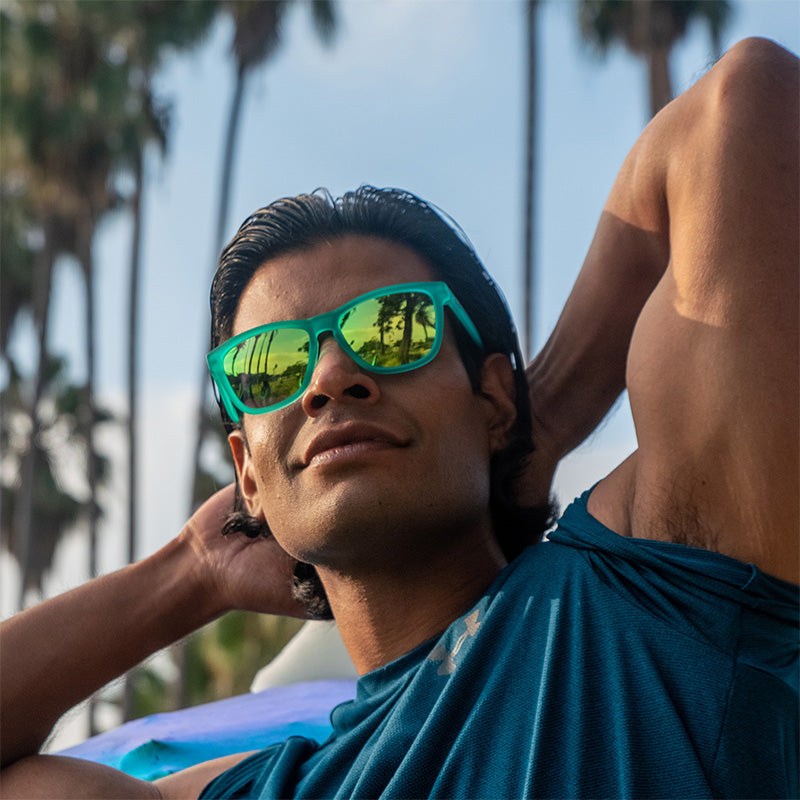 Een grijnzende gespierde man draagt een groenblauwe zonnebril met groenblauwe reflecterende glazen en buigt zijn biceps, palmbomen in de verte.