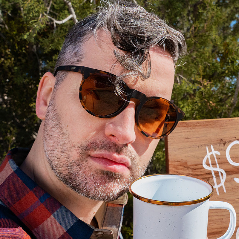 Een hipster barista kijkt veroordelend voor zich uit met een bruine zonnebril met schildpadglazen en bruine niet-reflecterende glazen.